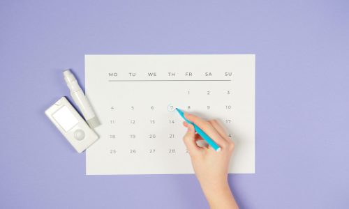 A hand marking a date on a calendar next to a glucometer on a purple background.